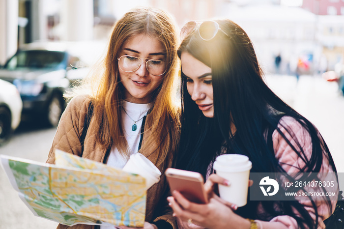 Two hipster girls discussing route of future travel sitting outdoors in urban setting