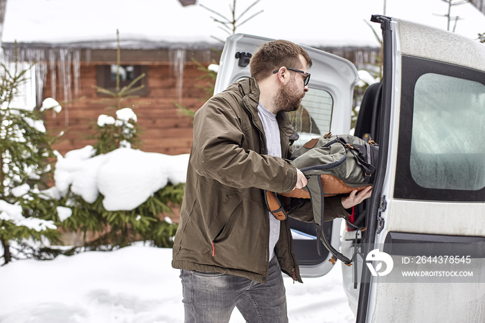 Bearded man in glasses puts things in the trunk of a car, against a background of snow-covered trees and a wooden house, on a winter day.