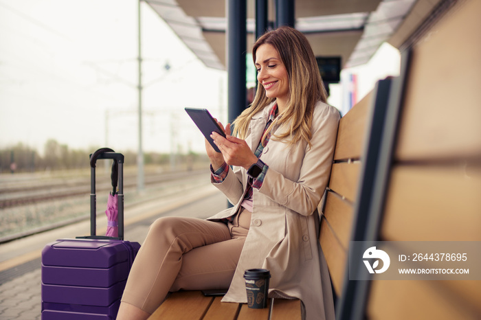 A young and beautiful woman with a suitcase is sitting at the train station waiting for a train while using a digital tablet for video calls and answering e-mails.