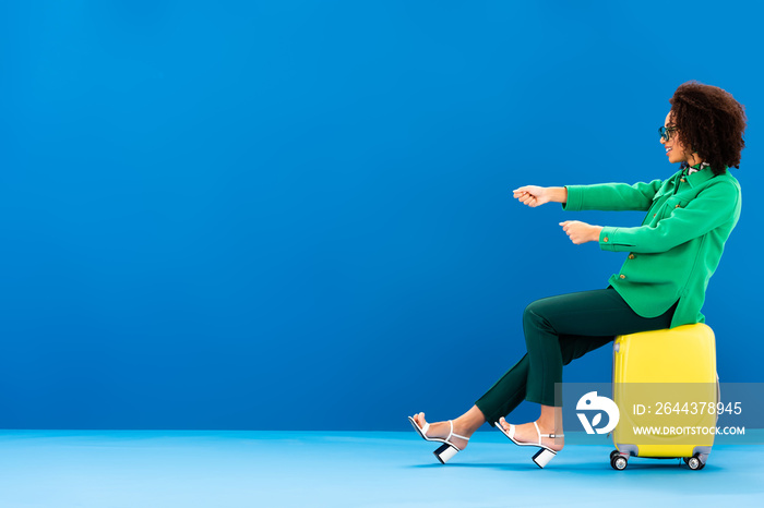 side view of smiling african american woman pulling and sitting on travel bag on blue background