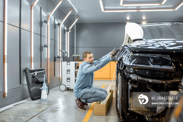 Man preparing protective film for sticking on a car body, marking it to with a pen at the vehicle service station. Concept of car body protection with special films