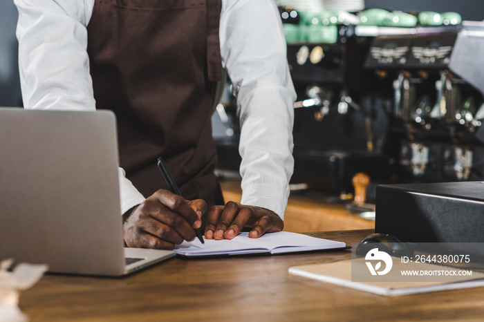 cropped shot of young african american barista taking notes and using laptop at coffee shop