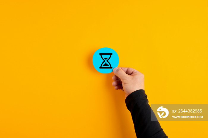 Male hand holds a blue badge with an hour glass icon on yellow background. Time management in business or project deadline