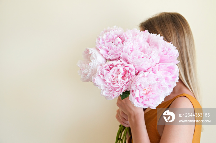 Conceptual portrait of young woman wearing mustard yellow dress covering her face with lush peonies. Female hiding behind beautiful bouquet of pink flowers. Close up, copy space, white background.