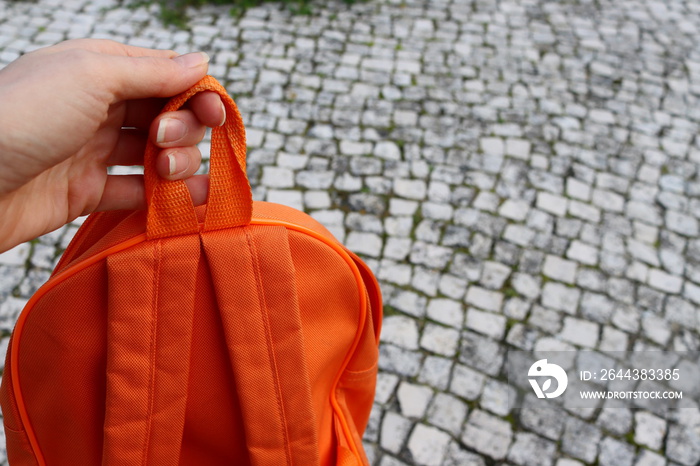 Female hand is holding an orange backpack on a gray background.