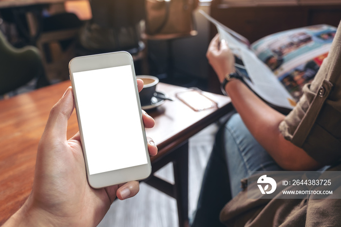 Mockup image of a hand holding white mobile phone with blank desktop screen and a woman reading magazine while sitting in cafe