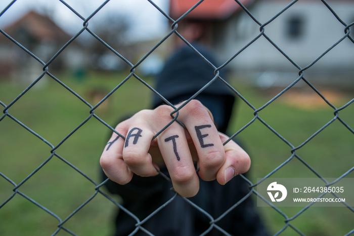 Aggressive teenage boy holding the wired fence at the correctional institute, the word hate is written on hes hand, conceptual image of juvenile delinquency .