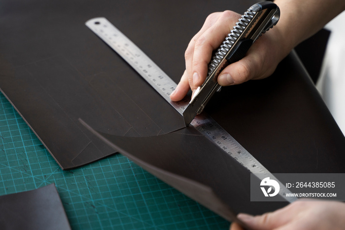 Men’s hand holding a stationery knife and metal ruler and cutting on a pieces for a leather wallet in his workshop. Working process with a brown natural leather. Craftsman holding a crafting tools.