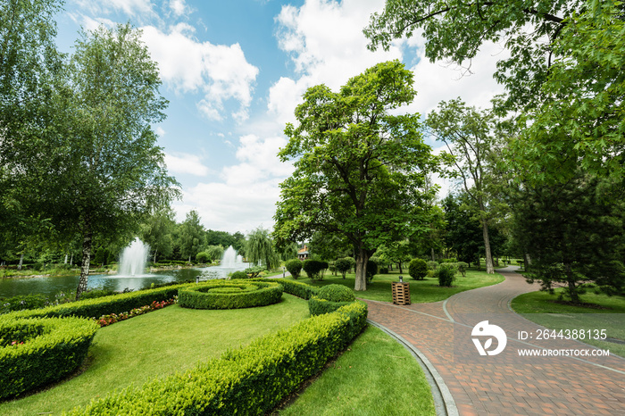 Walkway near green plants, trees and fountains against blue sky