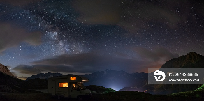 Panoramic night sky over the Alps. The Milky Way galaxy arc and stars over illuminated camper van. Camping freedom in unique landscape.