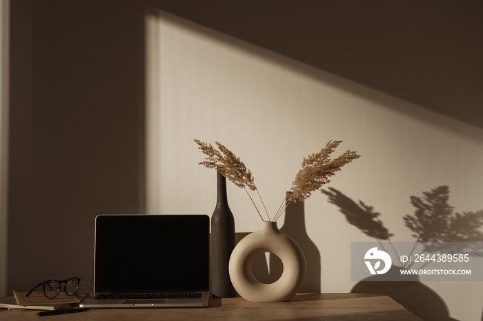 Laptop computer with blank screen on table with pampas grass bouquet in sunlight shadows on the wall. Aesthetic boho office workspace interior design. Sunlight shade reflections