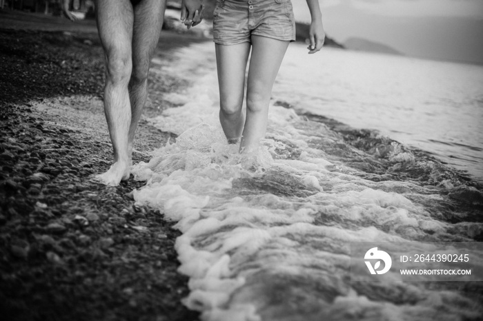 A black and white photo of the legs of a couple in love, in shorts and swimming trunks, walking along the ocean, with mountains in the background. The waves wash their feet.