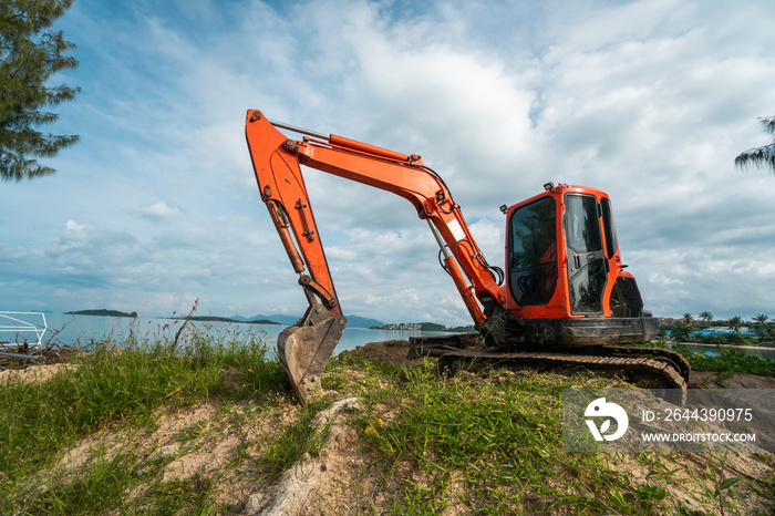 Small orange excavator on a ground against blue sky and sea for a works on construction site. Small tracked excavator standing on a ground with a blue sea on background. Heavy industry.