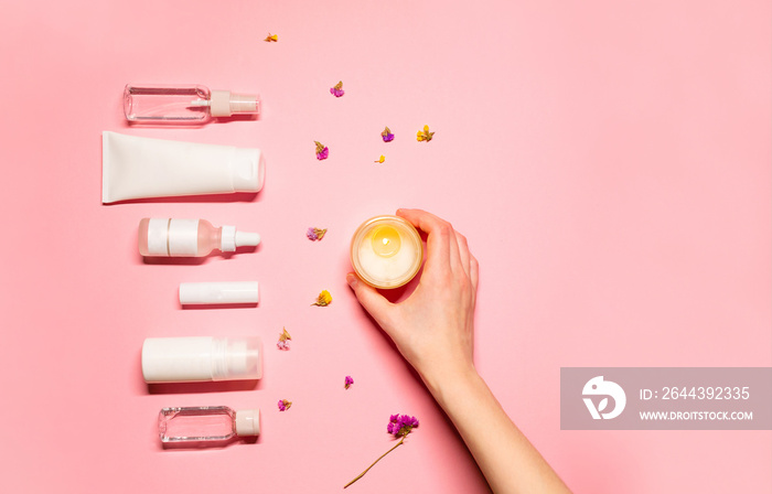 Flat lay photo with beauty products: lipstick, serum, hand cream, toner, facial spray; and a hand holding a scented candle on a pink background with colorful flower petals.