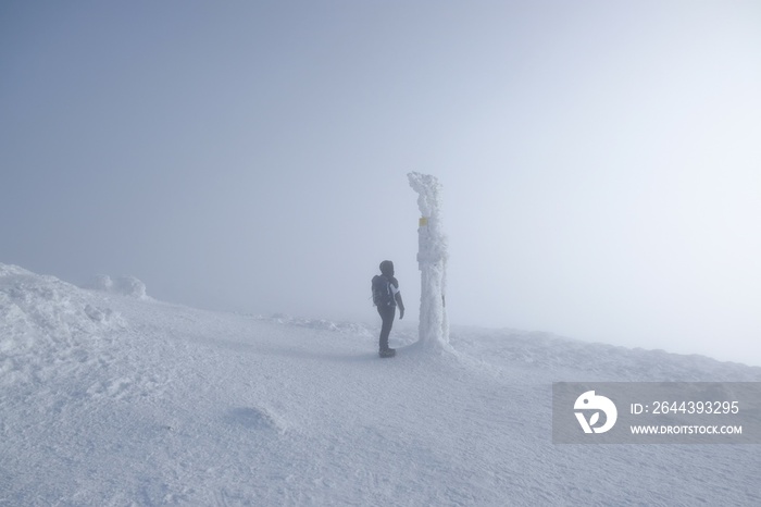 Woman standing next to frozen signpost on Babia Gora Peak on winter misty day. Diablak, Beskid Zywiecki, Poland