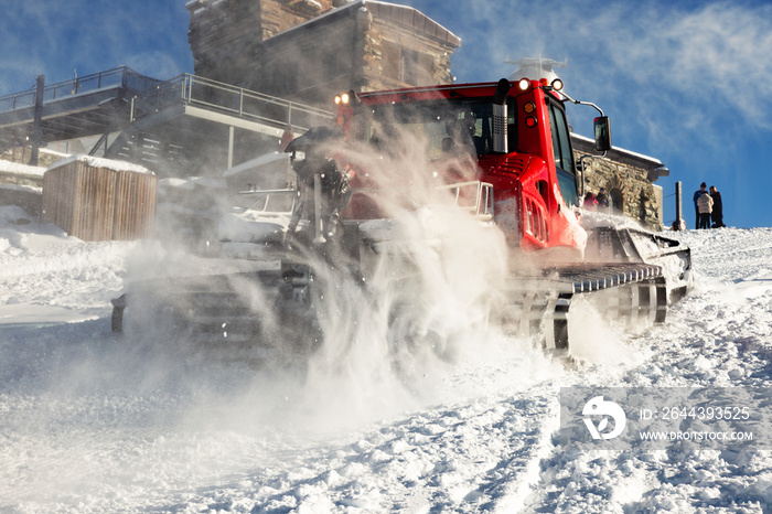 Photo of a red snowcat in action on the snowy track. Winter slope for skiing, during Christmas time. Snow dust rising behind the snowcat. Cable car station, and mountain refuge in the background