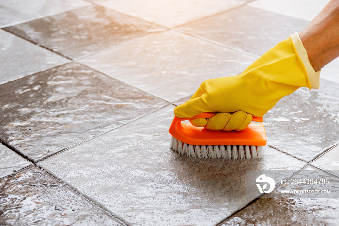 Hands wearing yellow rubber gloves are using a plastic floor scrubber to scrub the tile floor with a floor cleaner.