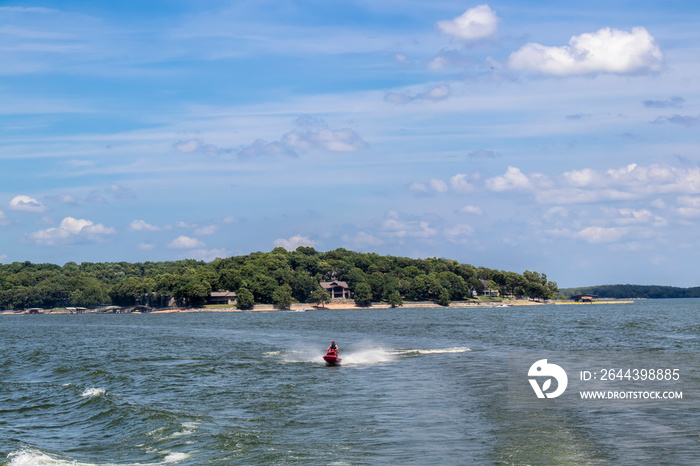 Person on red Personal Watercraft in choppy lake with homes and docks on shore and speedboat in background - Summer with cloudy blue cloud.