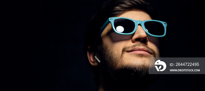 Close-up portrait of young guy with wireless earphones, looking up, wearing blue sunglasses, isolated on black background.