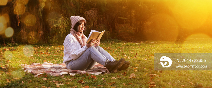 Young woman sitting on plaid and reading book in autumn park