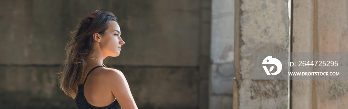 A thin athletic girl takes a break between classes on the background of the building in the early morning, enjoys silence and freedom. Panoramic image, toned.