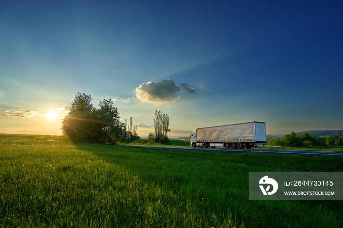 White truck driving on the road between green meadows in a rural landscape at sunset