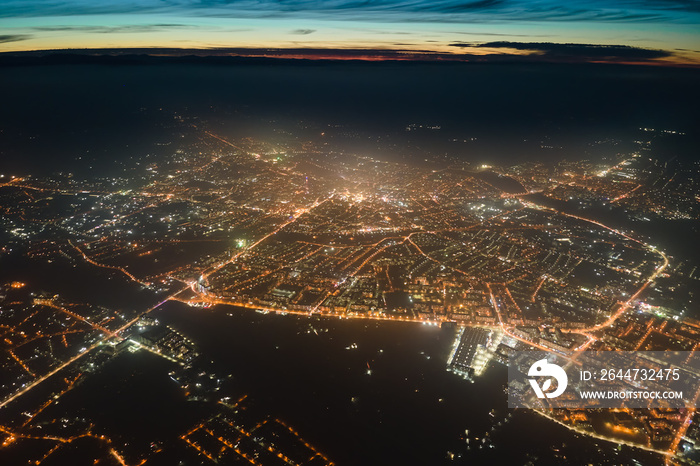 Aerial view from airplane window of buildings and bright illuminated streets in city residential area at night. Dark urban landscape at high altitude