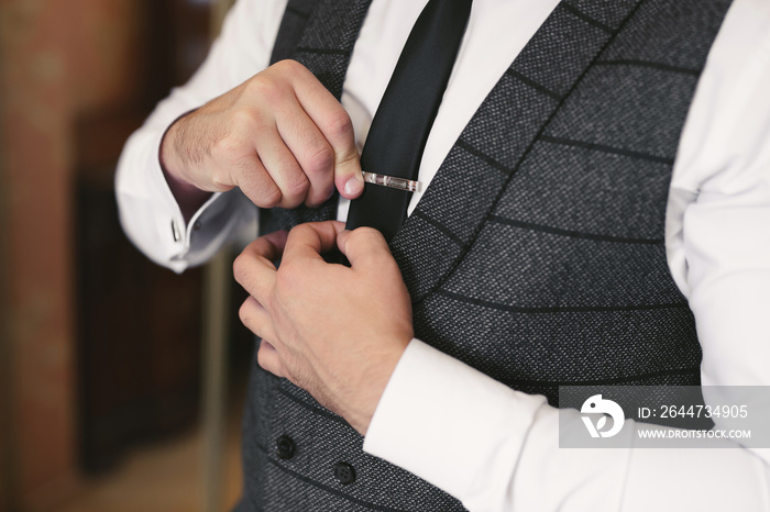 Elegant gorgeous groom getting ready in the morning in the hotel room, holding clip on his tie