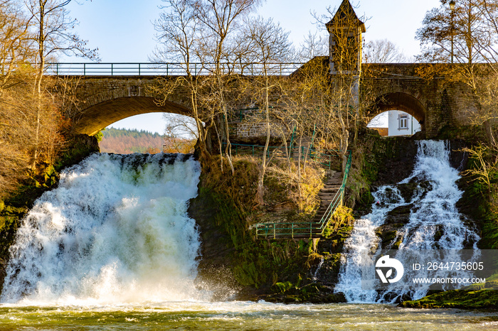 Rural stone vehicular bridge surrounded by leafless trees with a tower and two small waterfalls with abundant water flow on the Ambleve river, sunny day in the small town of Coo, Belgium