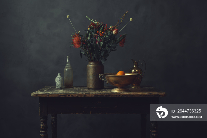 Old worn wooden table topped with a vase with flowers, a bronze bowl with oranges, a pitcher and two glass bottles.