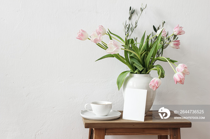 Rustic Easter still life scene. Blank greeting card mockup, cup of coffee and floral bouquet in white ceramic vase on wooden bench. Spring photo. Pink tulips flowers, olive tree branches on table.