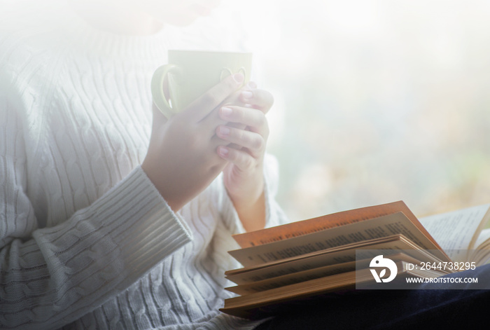 Girl is holding cup of tea and reading a book beside the window