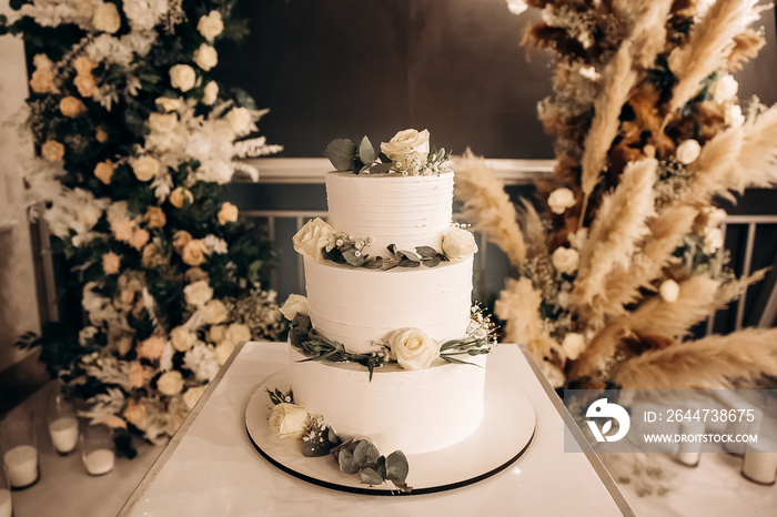 Large luxury wedding cake on a background of a flowering arch in the style of boho. Wedding dessert under the light of evening light bulbs. Wedding decor.