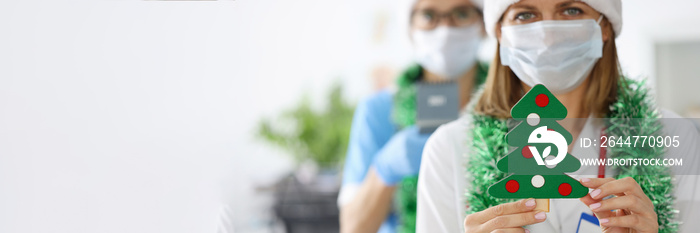 Doctors in santa hats and bright tinsel around their necks are holding toy christmas tree in clinic closeup