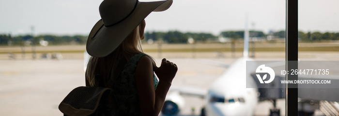 Travel tourist standing with luggage watching sunset at airport window. Unrecognizable woman looking at lounge looking at airplanes while waiting at boarding gate before departure. Travel lifestyle.