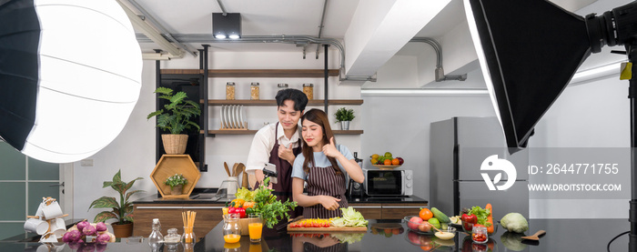 Asian couple spend time together in the kitchen. Young woman in apron cooking salad dish while his boyfriend recording vlog video for social blogger. Photography studio lights set up in the kitchen.