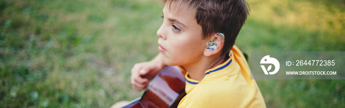 Hard of hearing preteen boy playing guitar outdoor. Child with hearing aids in ears playing music and singing song in a park. Hobby art activity for children kids. Web banner header.