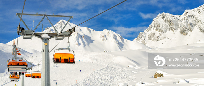 Ski lift ropeway on hilghland alpine mountain winter resort on bright sunny day. Ski chairlift cable way with people enjoy skiing and snowboarding.Banner panoramic wide view of downhill slopes