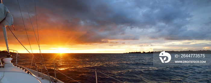 White yacht sailing in an open sea at sunset. A view from the deck to the bow, mast, sails. Epic cloudscape. Dramatic sky with glowing golden clouds after the storm. Racing, sport, leisure activity