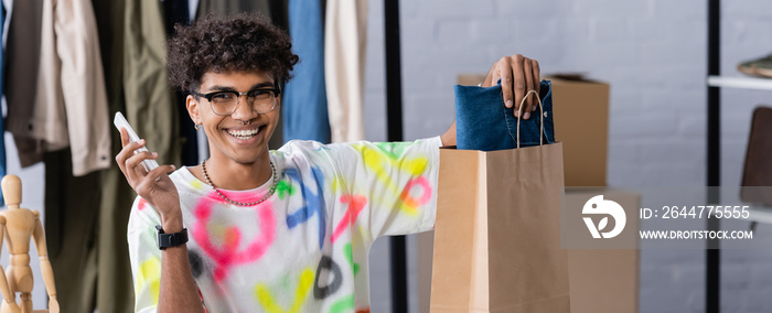 Cheerful african american small business owner with smartphone putting clothes in shopping bag, banner