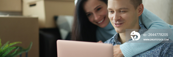 Young married couple communicating on social networks using laptop in apartment with boxes