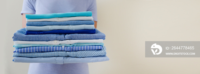 Cropped shot of young woman holding stack of perfectly folded shirts. Unrecognizable female with pile of different clothing in her hands. Laundry day concept. Close up, copy space, background