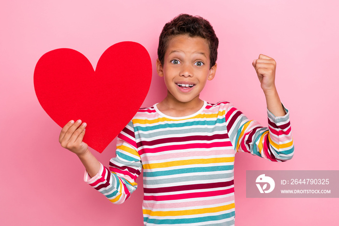 Portrait of adorable ecstatic boy with curly haircut dressed striped long sleeve hold red heart fist