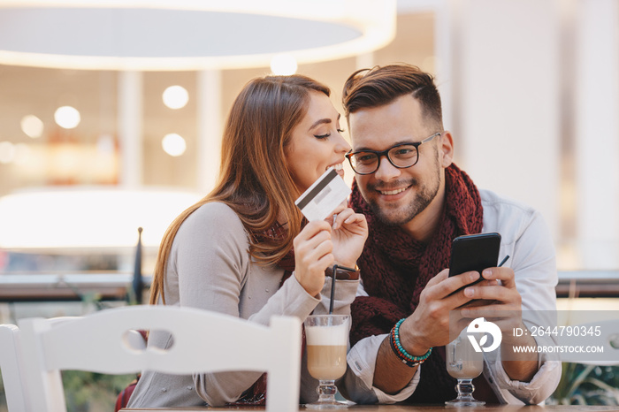 A couple with a credit card and a smartphone drinking coffee and shopping online at a cafe.