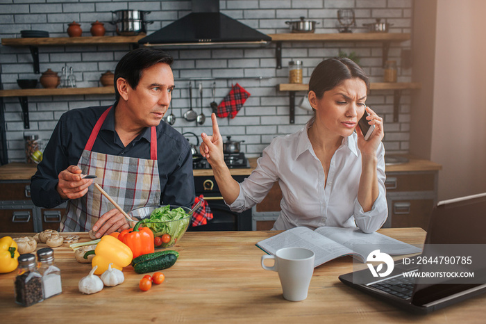 Busy woman sit at table and talk on phone. Man look and lean to her. They sit at table in kitchen. G