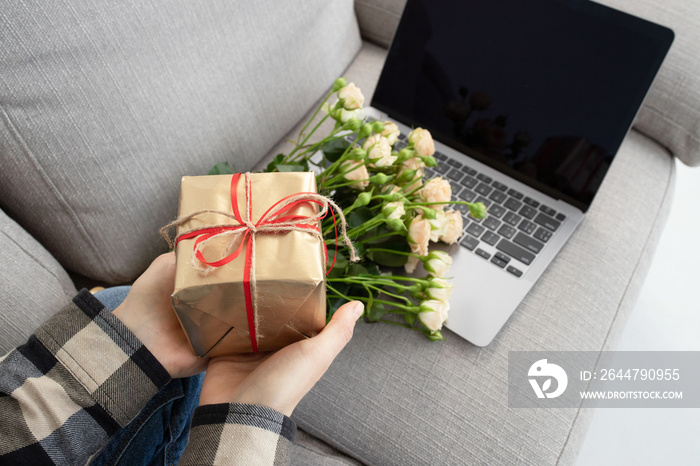 Gift box in female hands at the laptop screen. Beautiful bouquet of roses nearby. Concept of online 