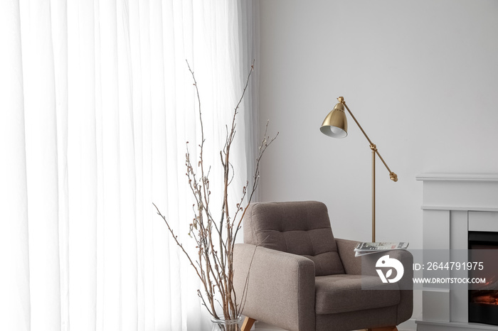 Vase with tree branches, armchair and lamp near light curtain in living room