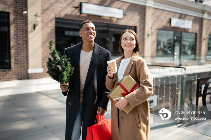 young woman with coffee to go and gift box smiling near stylish african american boyfriend with smal