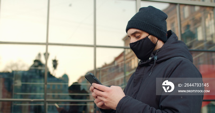 Side view of guy typing message while standing at street.Man with black cotton protective mask on fa