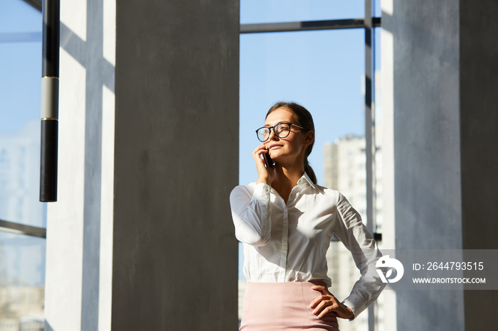 Content purposeful young business lady in glasses standing in modern corridor and talking by phone